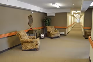 A long, well-lit hallway with beige walls and carpeted floor. Along the left side, there are two patterned armchairs, a small table with a plant and framed photos, and a large round mirror on the wall. The hallway extends into the distance with handrails on both sides.