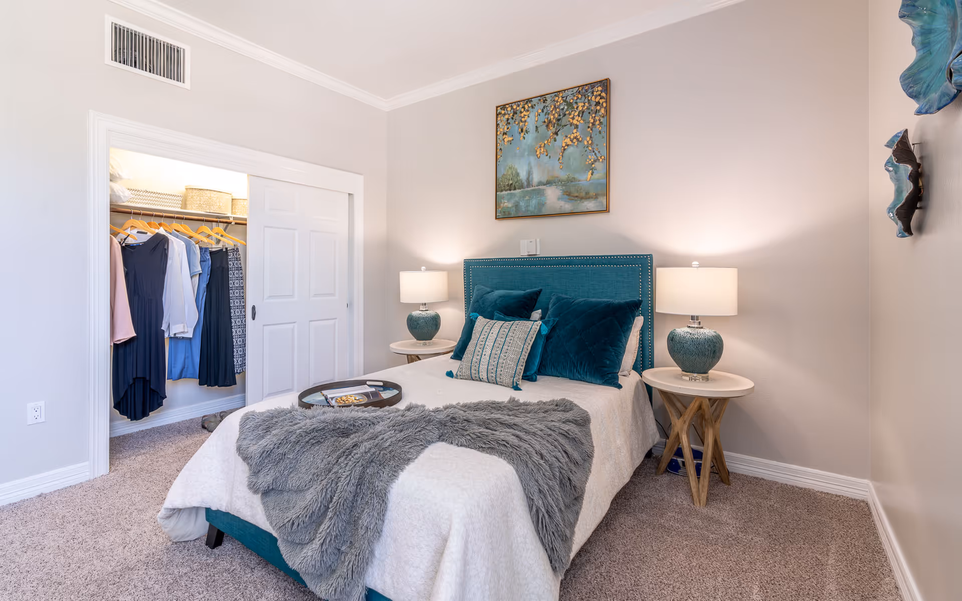 A cozy bedroom with a bed featuring teal and white bedding, two bedside tables with matching lamps, a gray throw blanket, and a closet with clothes hanging inside. The walls are light-colored with decorative artwork above the bed and on the side wall.