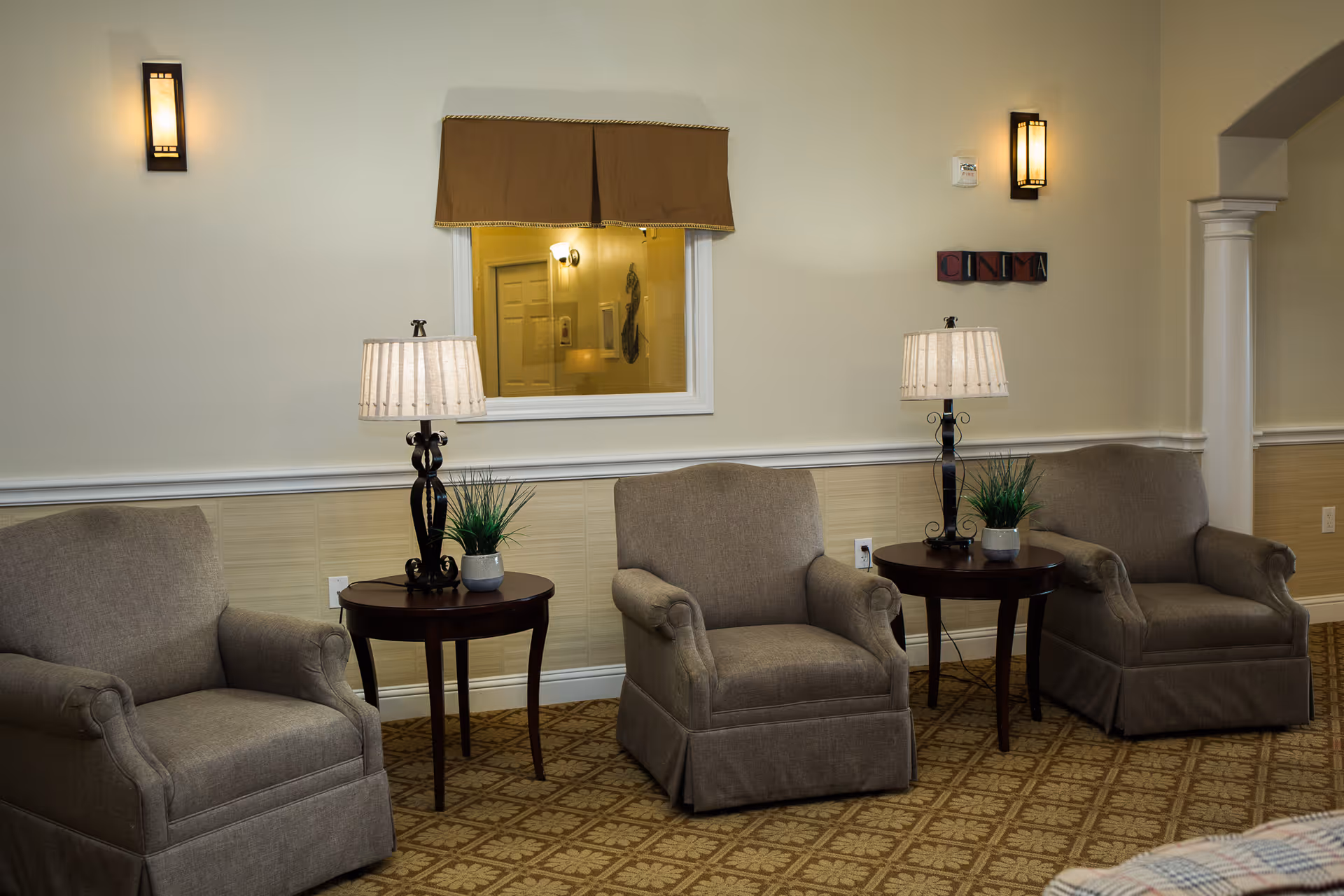 A beige lounge with three upholstered armchairs, two round side tables topped with lamps and small plants, and wall sconces.