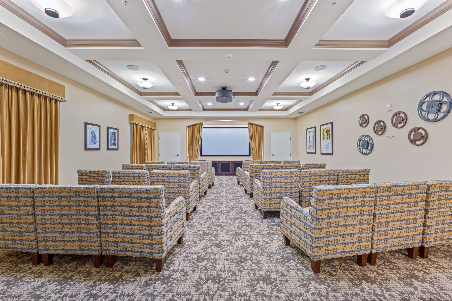 Interior view of a small theater or screening room with multiple rows of patterned armchairs facing a large projection screen. The room has a coffered ceiling with recessed lighting, beige walls decorated with framed pictures and film reel wall art, and gold curtains on the windows.