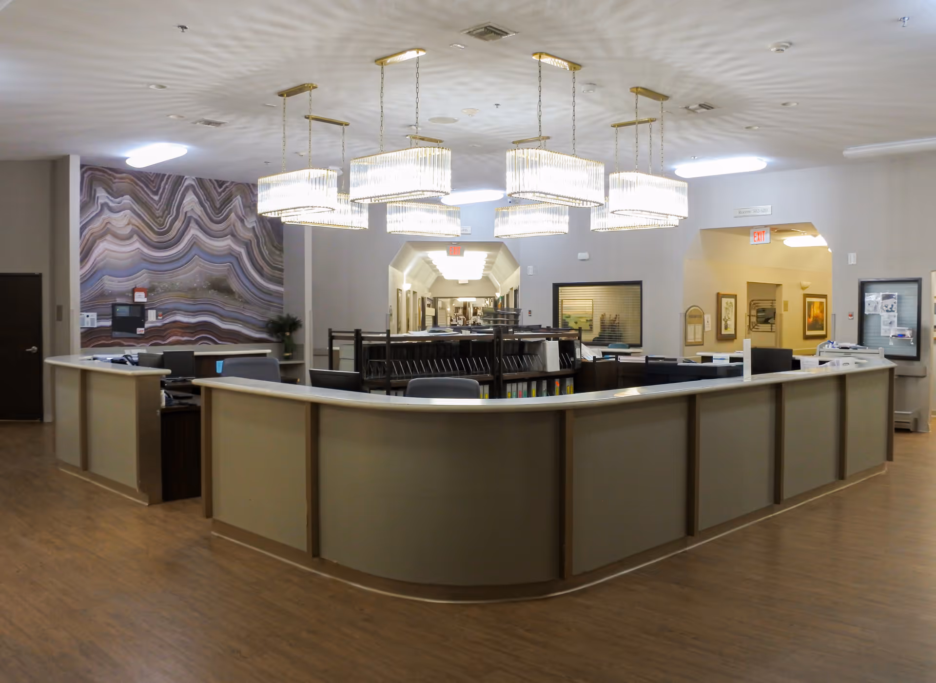 Interior view of a reception or nurse station area in a healthcare facility with a large curved counter, several hanging rectangular chandeliers, wood flooring, and a decorative wall with a wavy pattern on the left side.