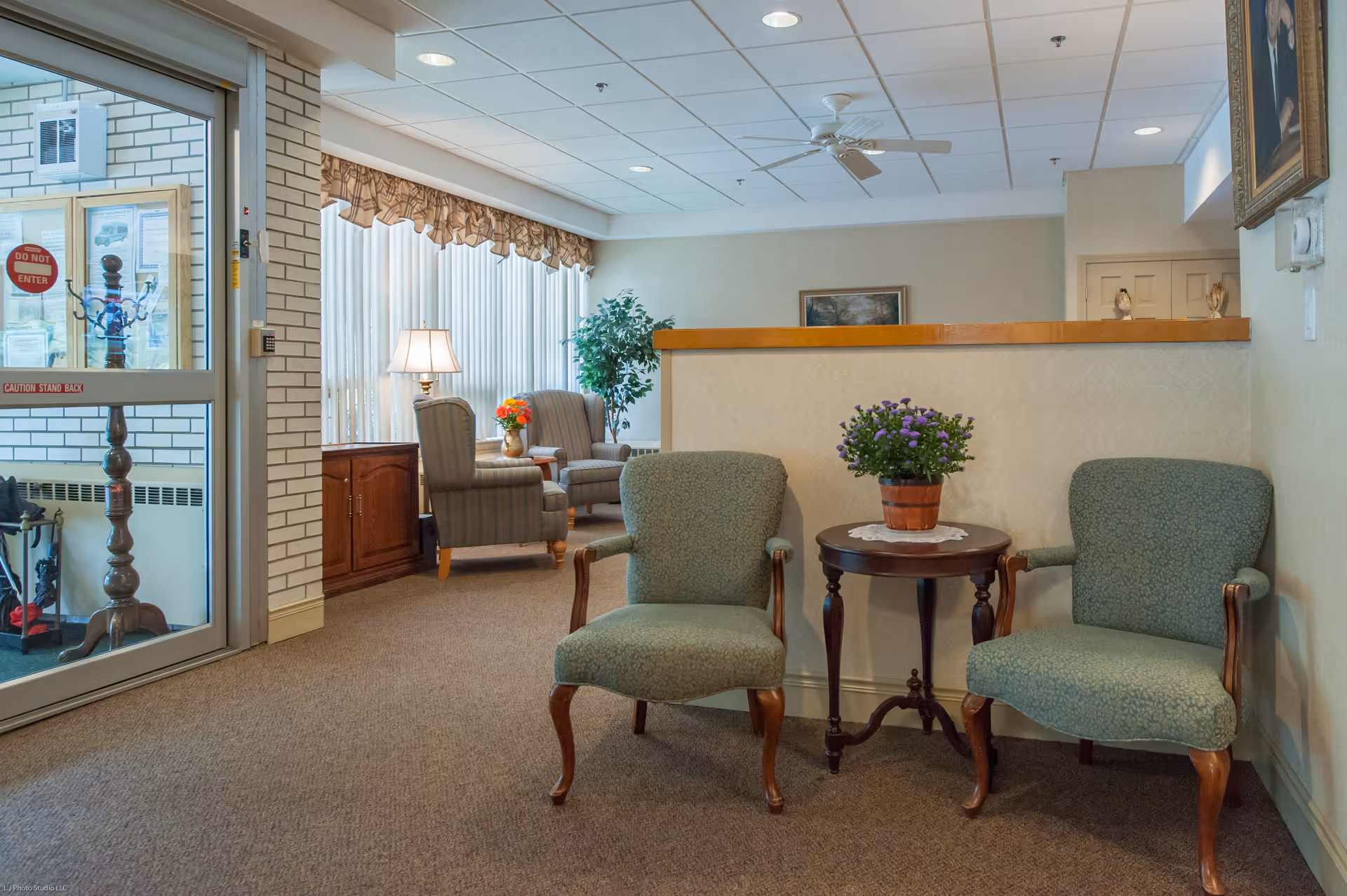 A cozy waiting area in a senior living facility with two green upholstered armchairs and a small round wooden table with a potted purple flower. In the background, there are two more armchairs near a window with vertical blinds and a table lamp. The entrance door has a 'Do Not Enter' sign and a caution sticker. The walls are light-colored, and the carpet is brown.