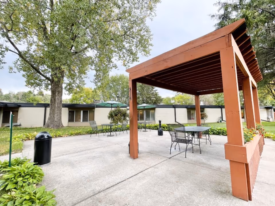 Outdoor patio area at The Villas at Roseville featuring a wooden pergola with a table and chairs underneath, additional tables and chairs with green umbrellas, surrounded by green grass, trees, and a single-story building in the background.