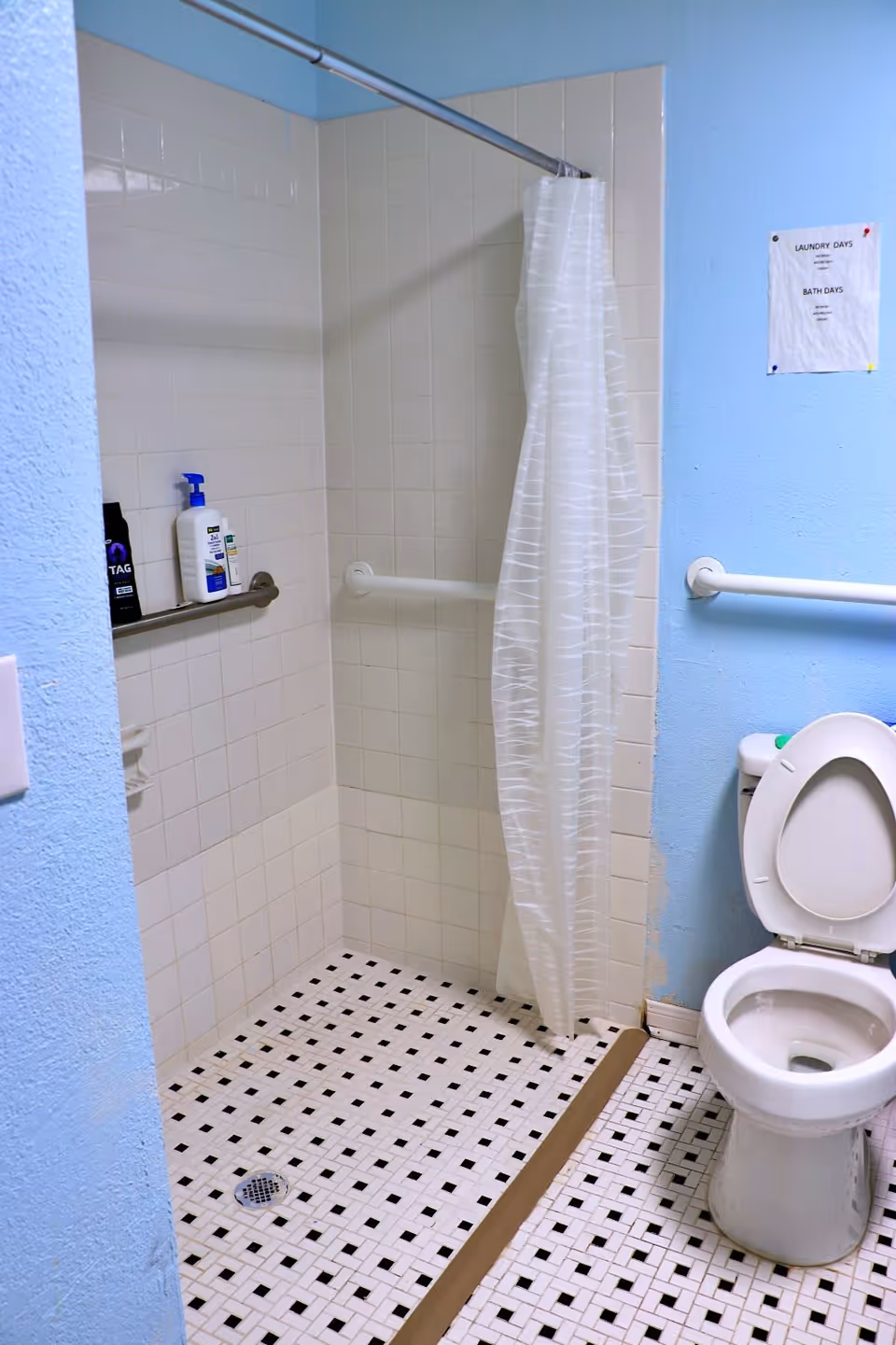 A bathroom with a tiled walk-in shower featuring a white curtain and grab bars, a toilet with the lid open, and a blue wall with a small sign posted. The floor has a black and white checkered tile pattern.
