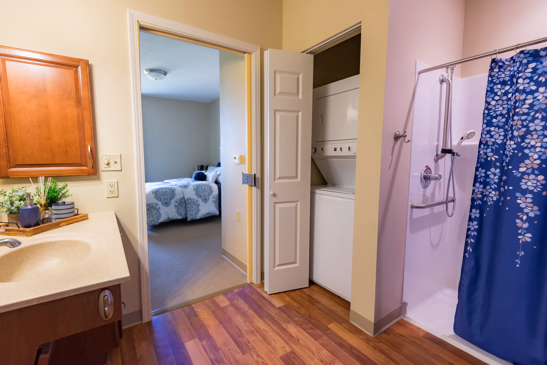 Interior view of a senior living facility bathroom with a sink and countertop on the left, a shower with a blue floral curtain on the right, and a stacked washer and dryer unit in a closet with bi-fold doors. Through an open doorway, a bedroom with a bed covered in white and blue patterned bedding is visible.