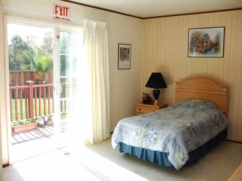 A bright bedroom with a single bed covered in a floral bedspread, a wooden nightstand with a black lamp, framed pictures on the wall, and a sliding glass door leading to an outdoor deck with plants visible outside.
