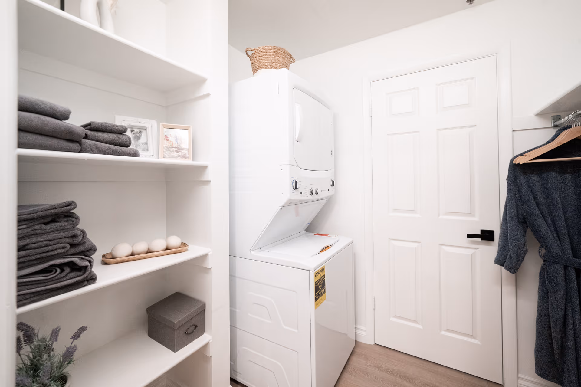 Small laundry room with a stacked washer-dryer, white built-in shelves holding folded towels and decor, and a hanging robe by a closed door.