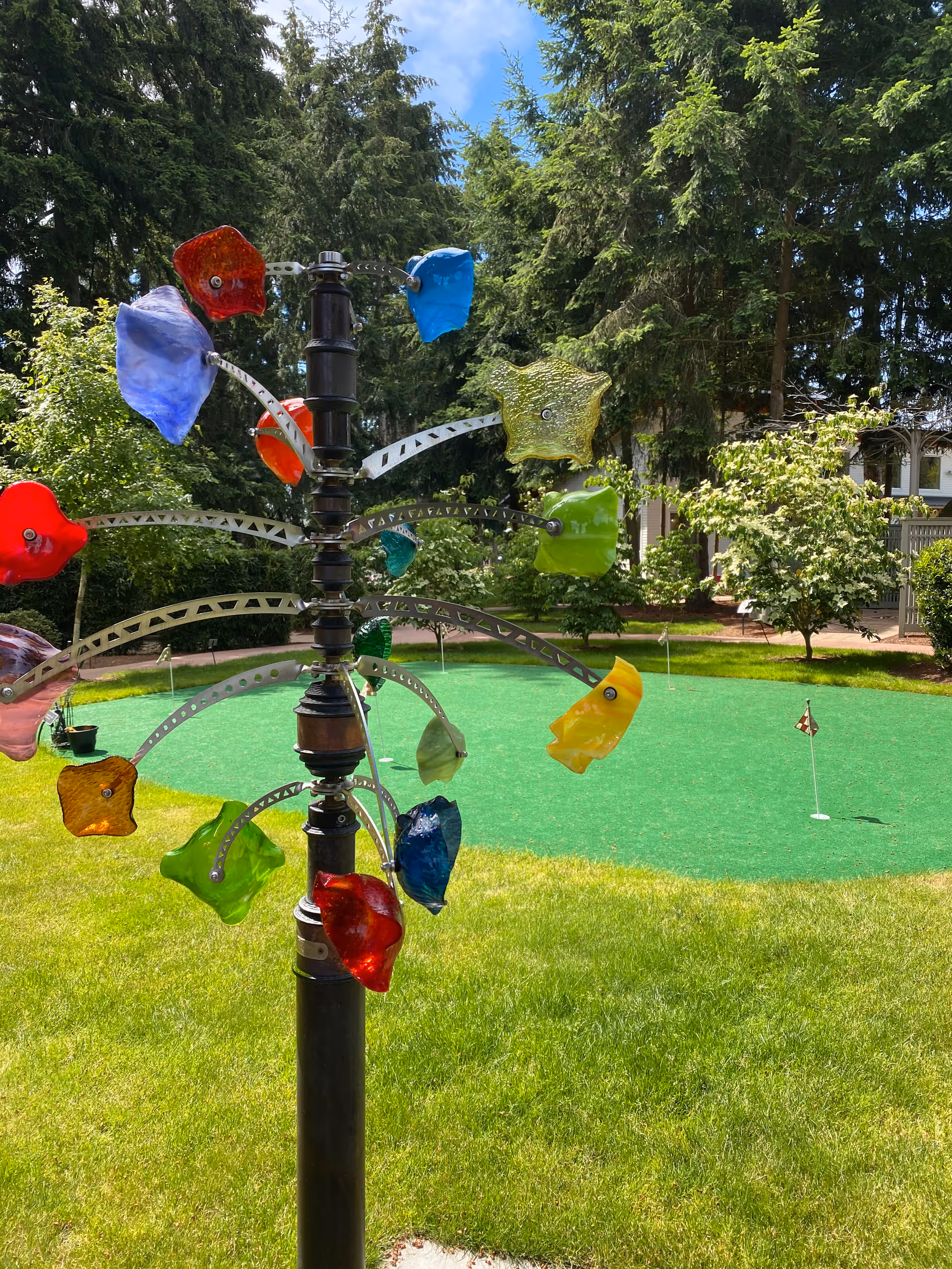 A colorful outdoor kinetic sculpture with various translucent, flower-shaped pieces in red, blue, green, yellow, and orange mounted on metal arms attached to a central black pole. The sculpture is set in a grassy yard with a putting green and small golf flags in the background, surrounded by trees and shrubs under a partly cloudy sky.
