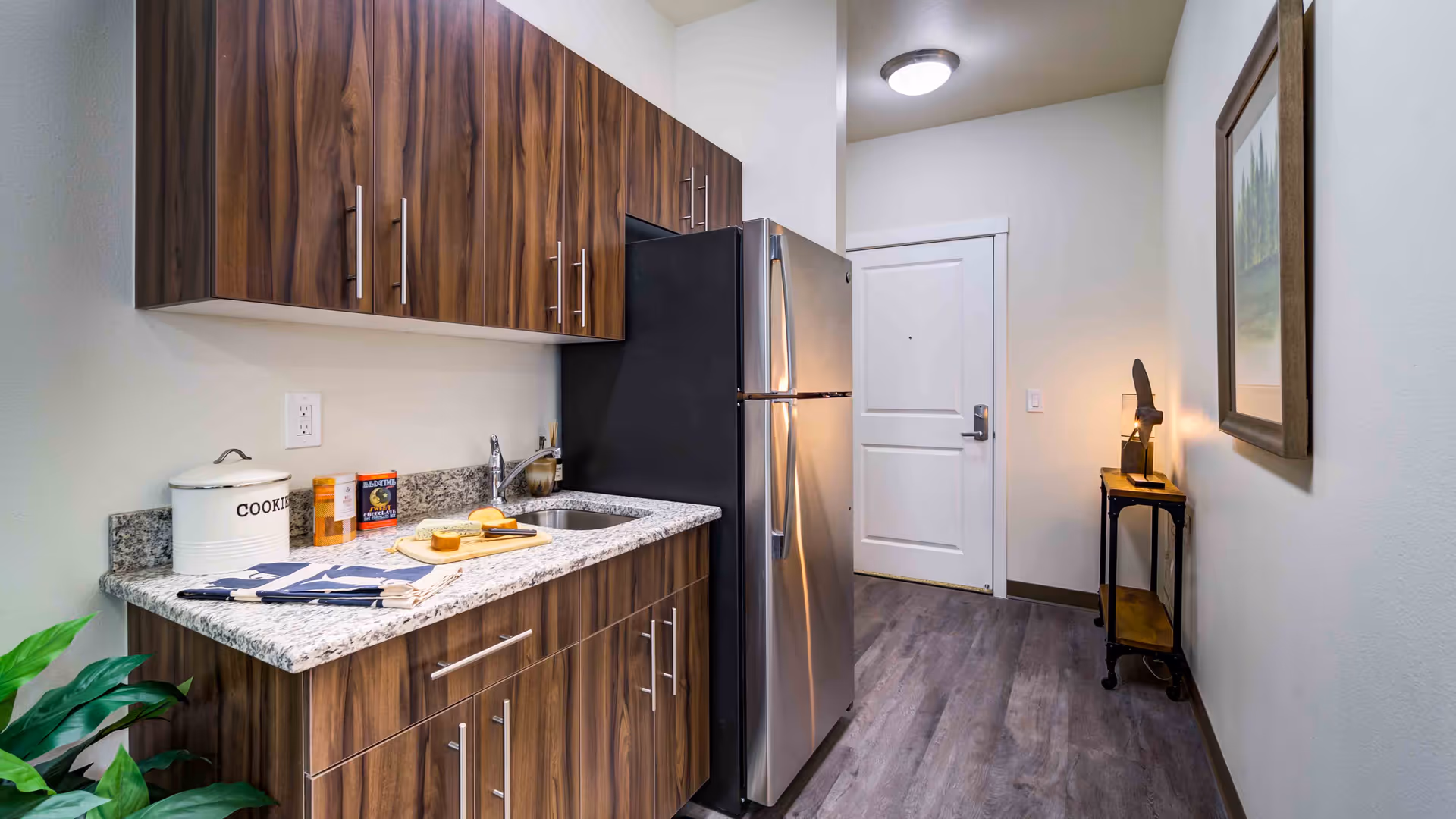 A modern kitchen area with wood grain cabinets, a granite countertop, a stainless steel refrigerator, a small sink, and various kitchen items including a cookie jar and a cutting board with bread. The kitchen is adjacent to a hallway with a white door, a small table with a lamp, and a framed picture on the wall.