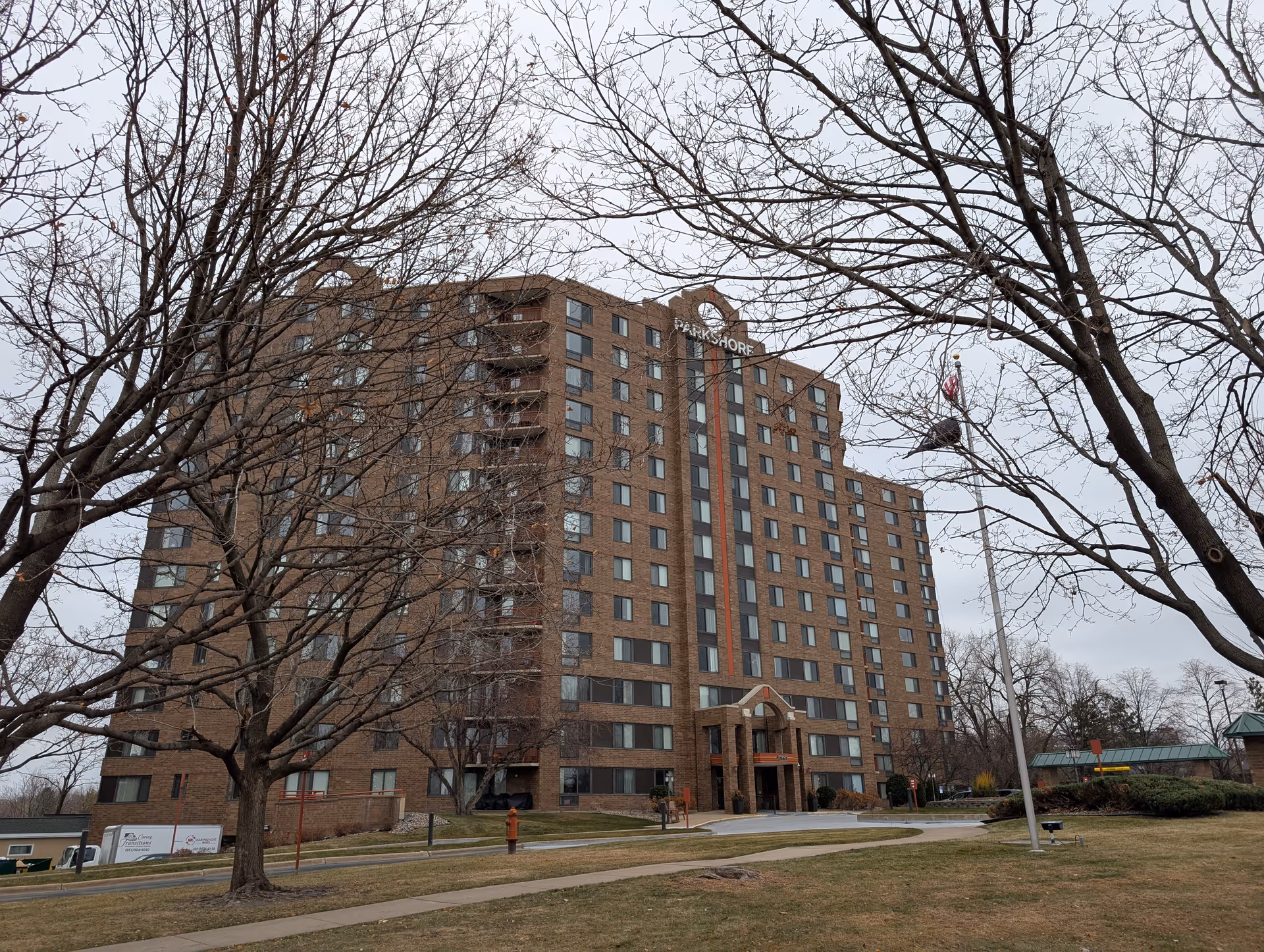 Exterior view of a multi-story brick building named Parkshore Senior Campus with leafless trees in the foreground and a flagpole with an American flag on the right side.