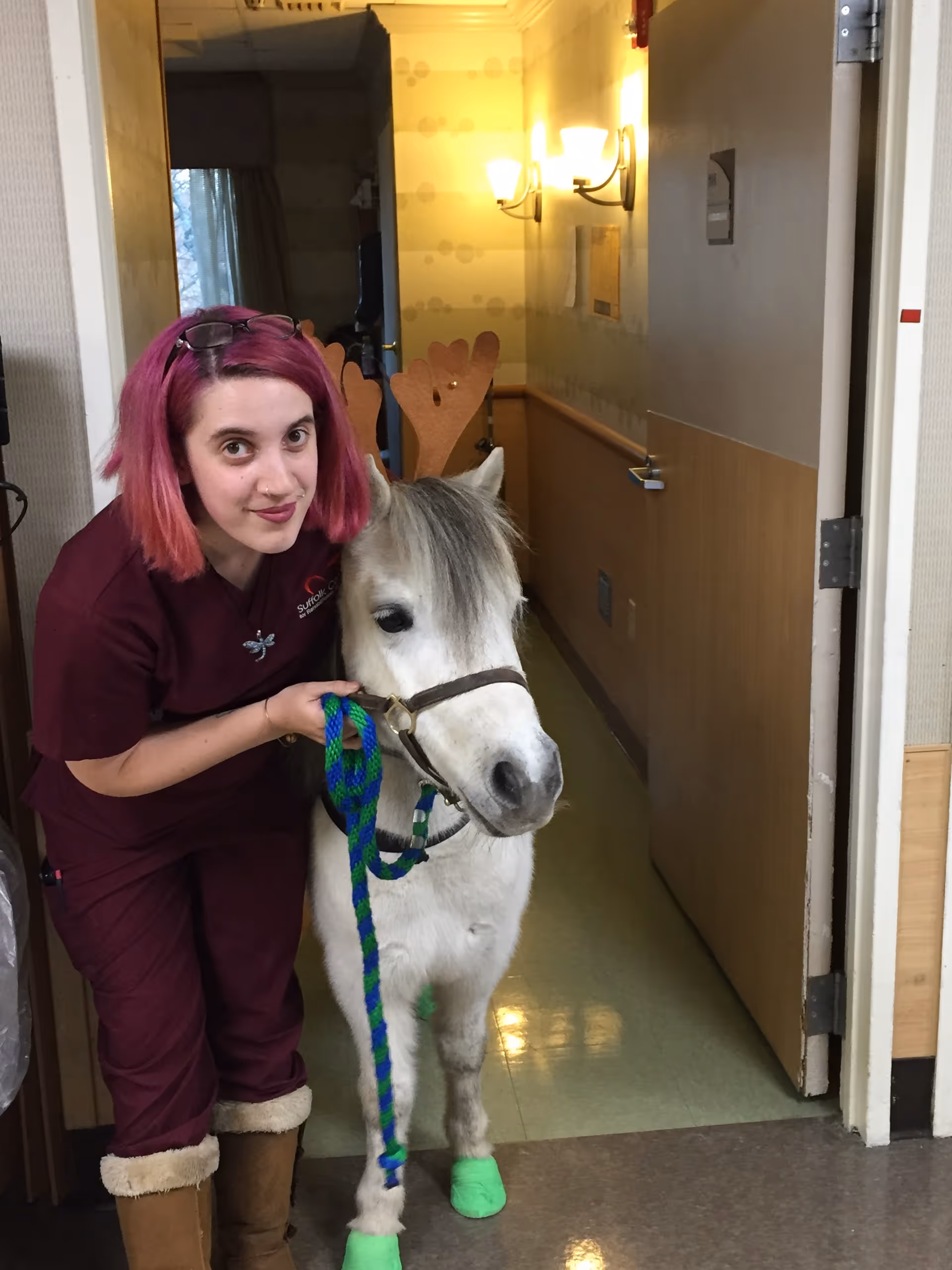 A person with pink hair in scrubs poses in a hallway holding a small white pony wearing antler headband and green hoof wraps.