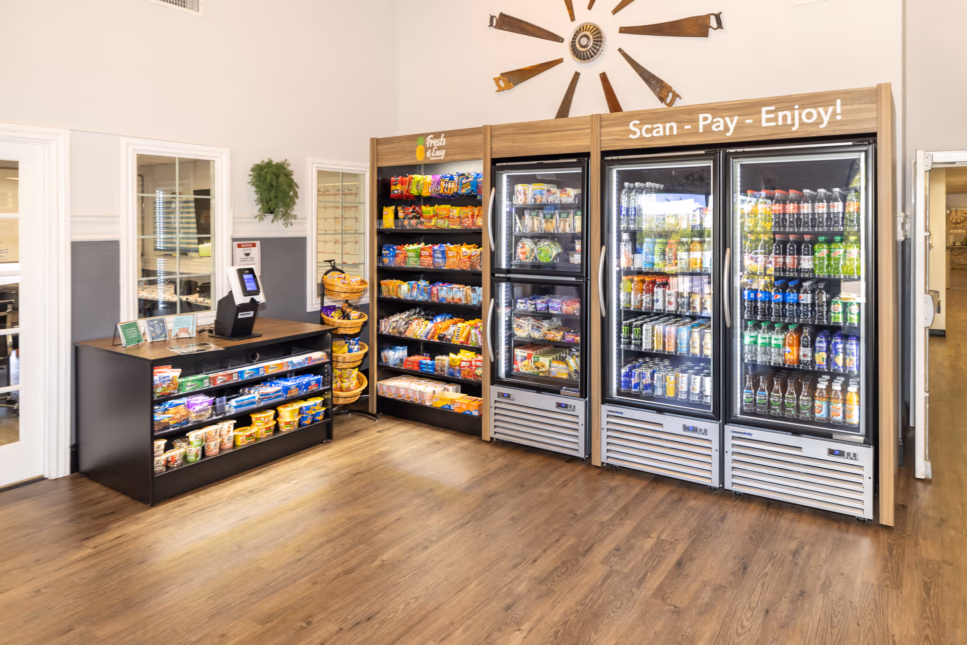 A small convenience area inside a facility with shelves stocked with snacks and refrigerated coolers filled with drinks. There is a self-checkout kiosk on a counter to the left, and a decorative wall piece made of saw blades above the coolers. The floor is wooden and the walls are painted white and gray.