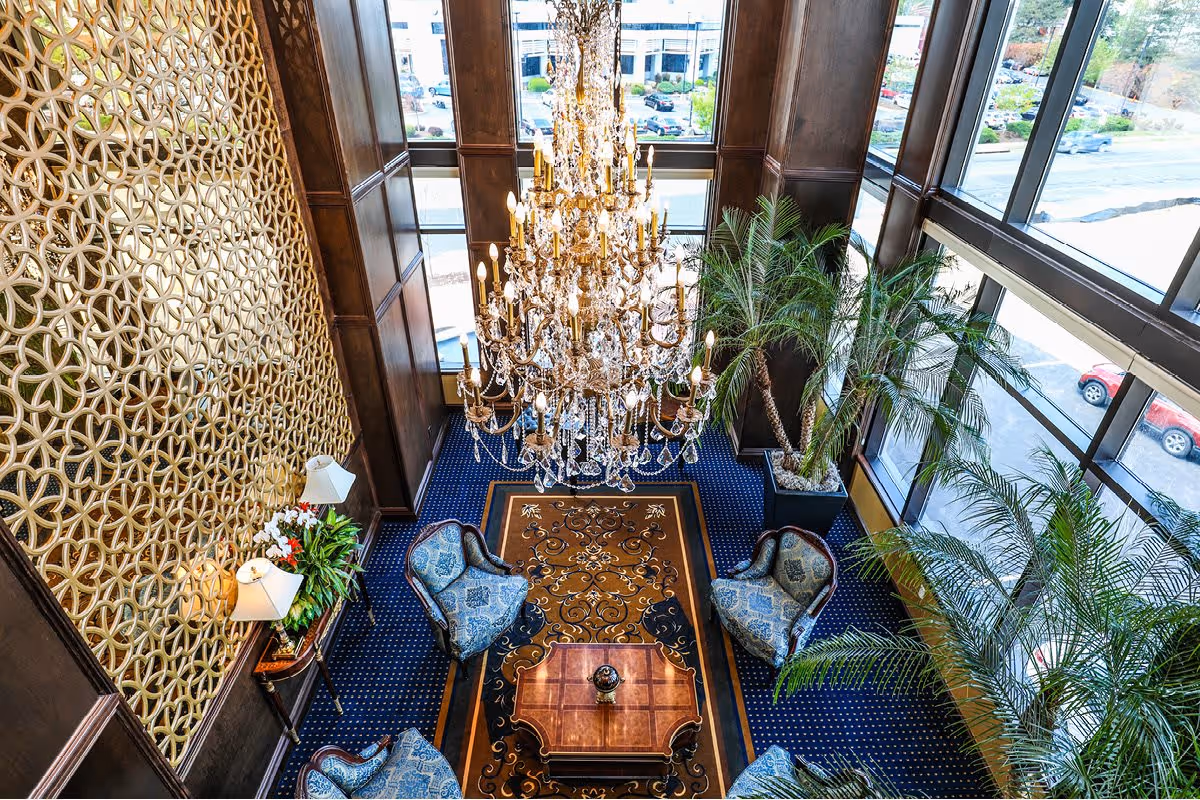 View from above of an elegant lobby area with a large crystal chandelier hanging from the ceiling, a patterned carpet, four upholstered chairs around a wooden coffee table, tall potted palm plants, and large windows letting in natural light.