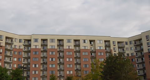 Exterior view of a multi-story senior living facility building with beige and reddish-brown facade, multiple balconies, and windows under a cloudy sky. Trees and greenery are visible in the foreground.