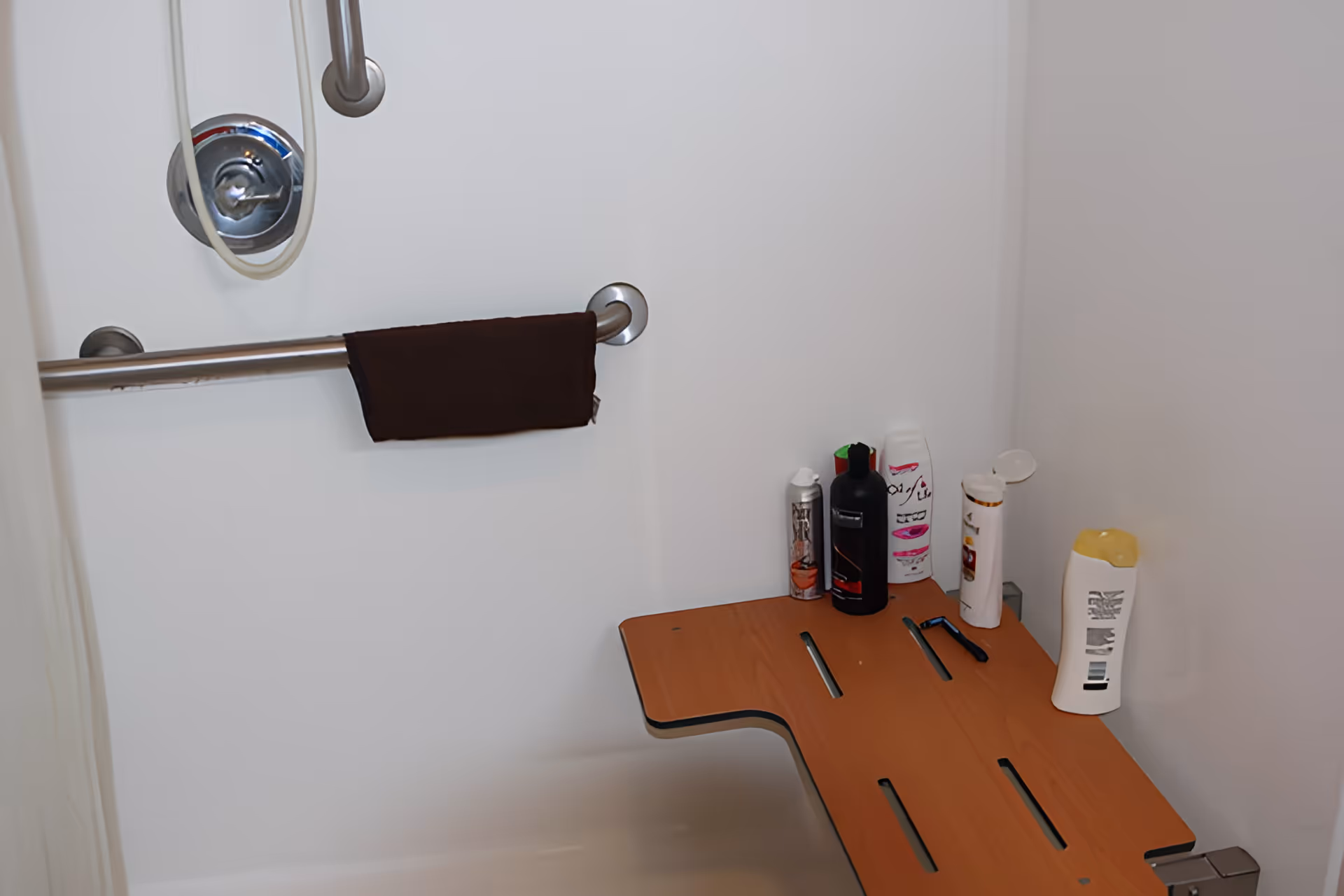 A shower area with a metal grab bar holding a dark brown towel, a shower control knob, and a wooden shower seat with various bottles of shampoo, conditioner, and body wash placed on it.