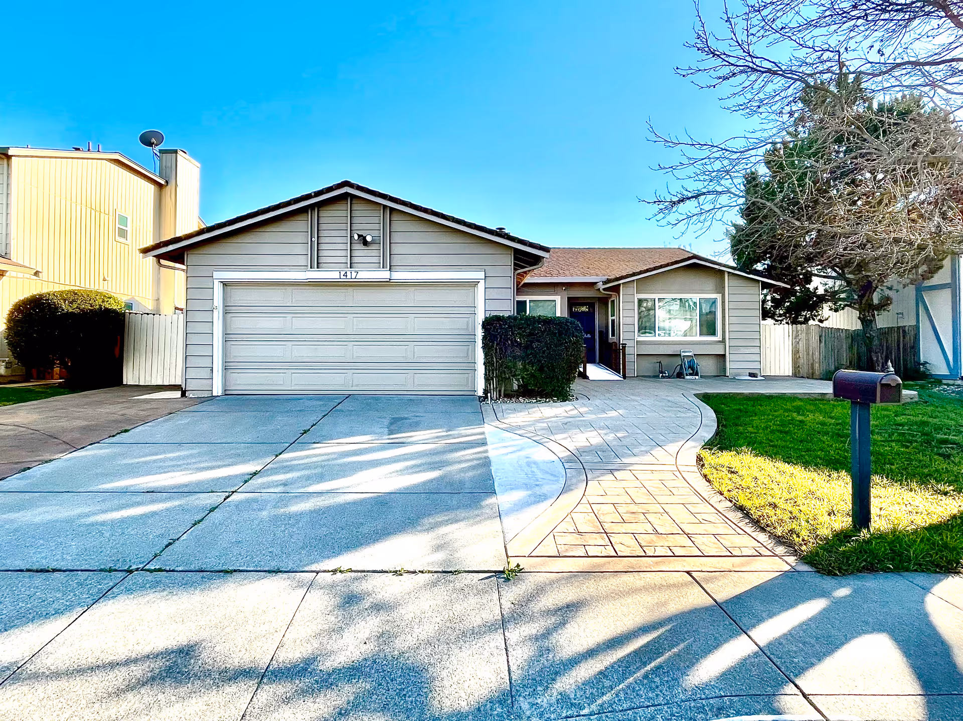 Front exterior view of a single-story house with a two-car garage, a curved paved walkway leading to the front door, a mailbox on the right side, and a tree with no leaves on the right lawn under a clear blue sky.