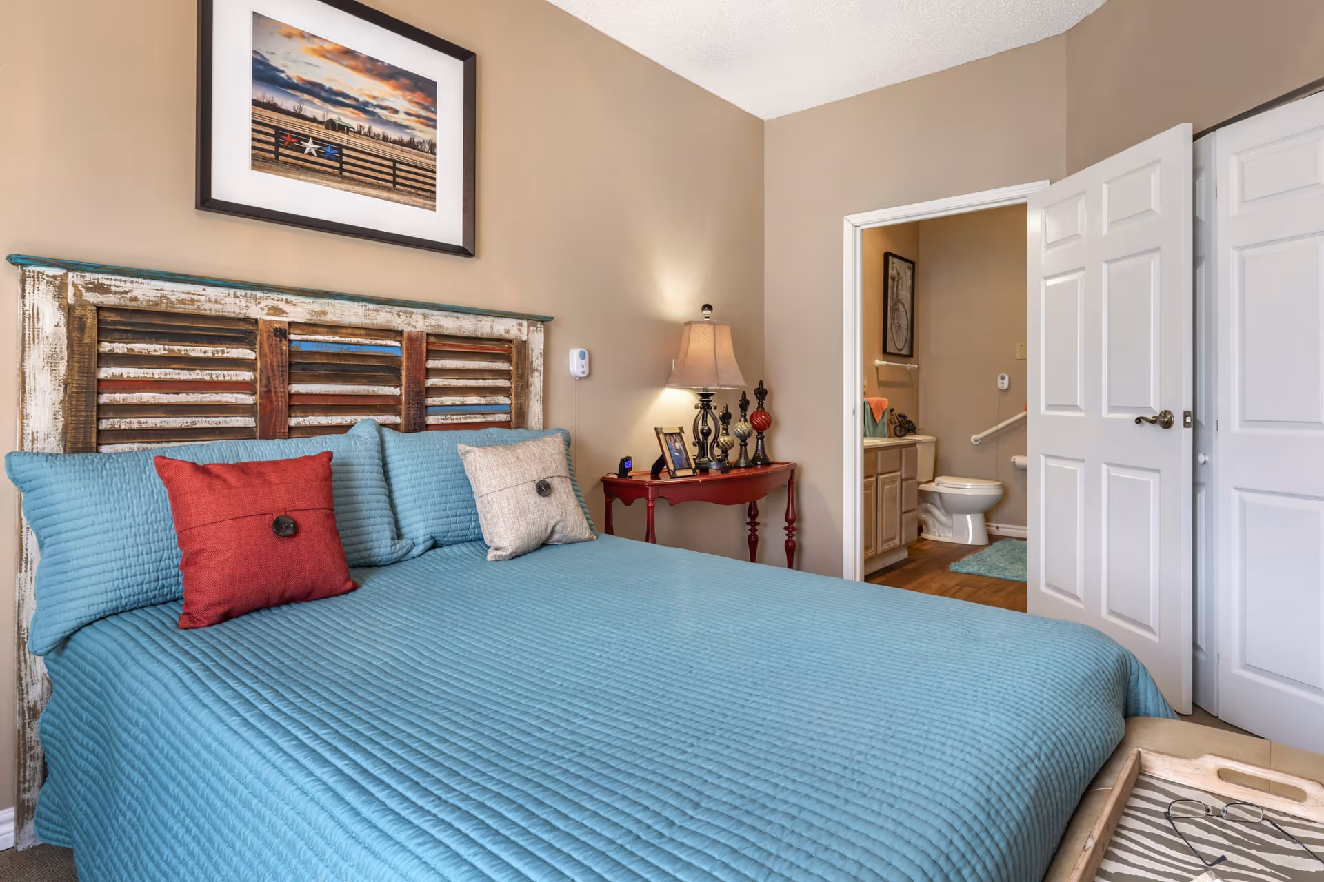 A bedroom with a bed covered in a blue quilt and pillows, including a red and a beige one. The bed has a rustic wooden headboard. A framed picture hangs above the bed. Next to the bed is a red side table with a lamp, decorative items, and a photo frame. An open door reveals a bathroom with a toilet, sink, and grab bars.