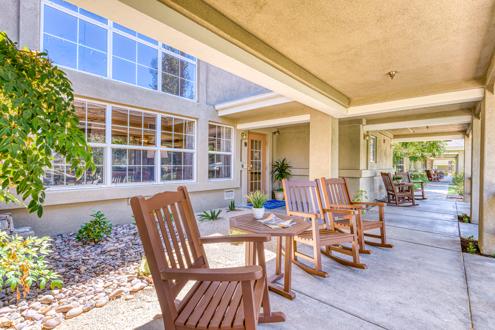 Covered outdoor walkway at a senior living facility with several wooden rocking chairs and small tables. The walkway is adjacent to a building with large windows and some landscaping with rocks and plants.