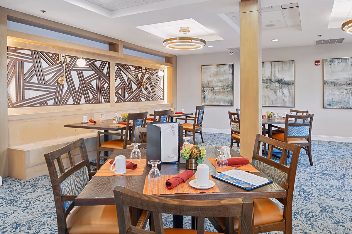 A bright and modern dining room with wooden tables and chairs, each table set with placemats, napkins, cups, glasses, and menus. The room features decorative wall art, a patterned carpet, and a wooden partition with geometric designs. Ceiling lights provide soft illumination.