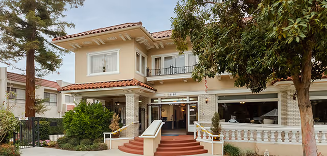 Exterior front view of a two-story building with beige walls and a red tile roof, featuring a wide entrance with steps and handrails, surrounded by trees and greenery.