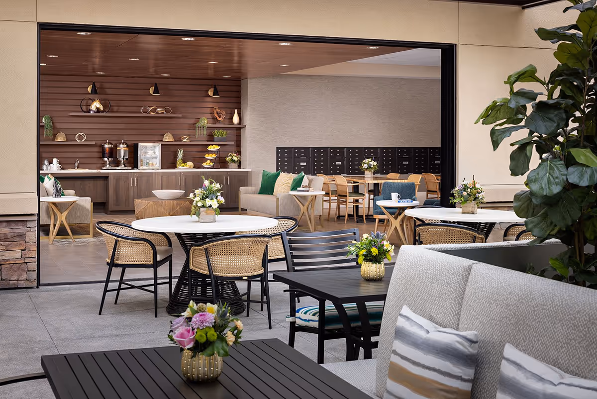 Outdoor patio area with tables and chairs, decorated with small flower arrangements. The patio opens into an indoor lounge area with sofas, chairs, tables, and a wooden wall with shelves holding decorative items and plants.
