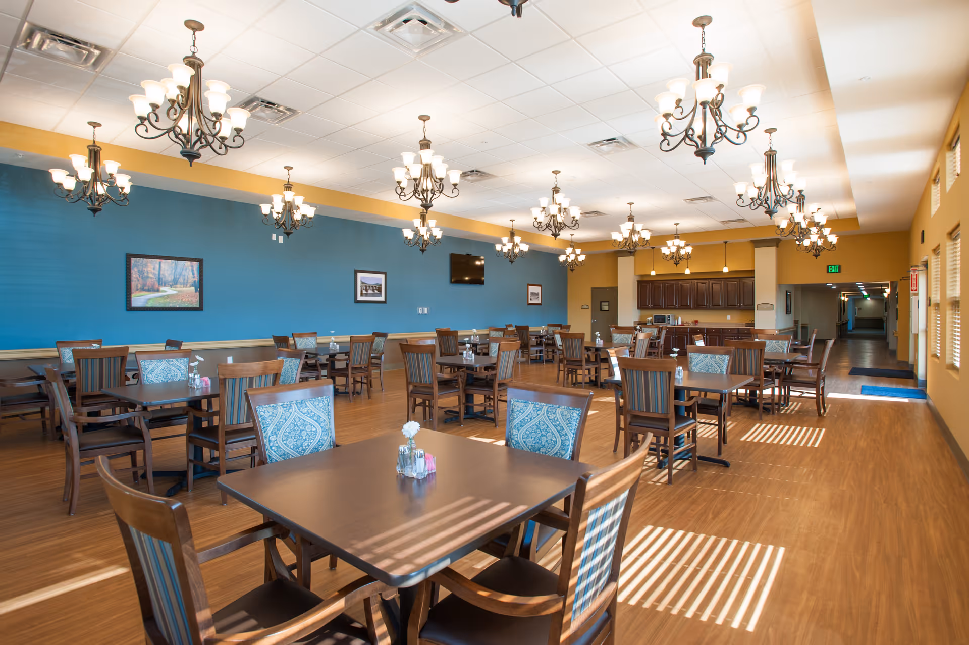 A spacious dining room with multiple wooden tables and chairs, some with patterned cushions. The room features a blue accent wall with framed pictures, several ornate chandeliers hanging from a white ceiling, and a wooden floor. There is a kitchen area with dark cabinets and a hallway leading to other parts of the facility.