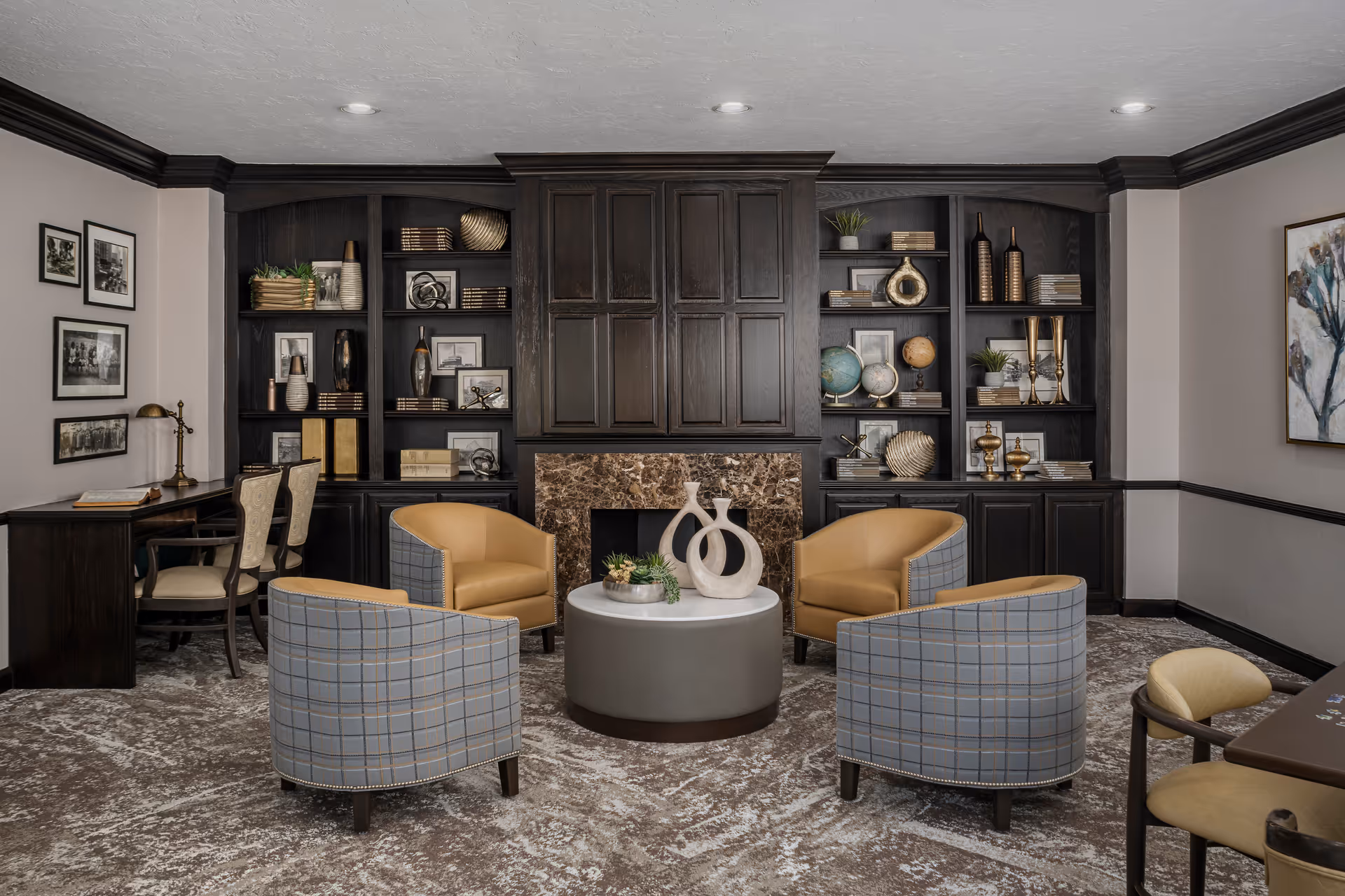 A cozy living room area with four armchairs arranged around a round coffee table. The chairs are a mix of tan leather and blue plaid fabric. Behind the seating area is a dark wood built-in bookshelf with various decorative items and books, and a marble fireplace in the center. The room also features a desk with two chairs on the left side and framed artwork on the walls.