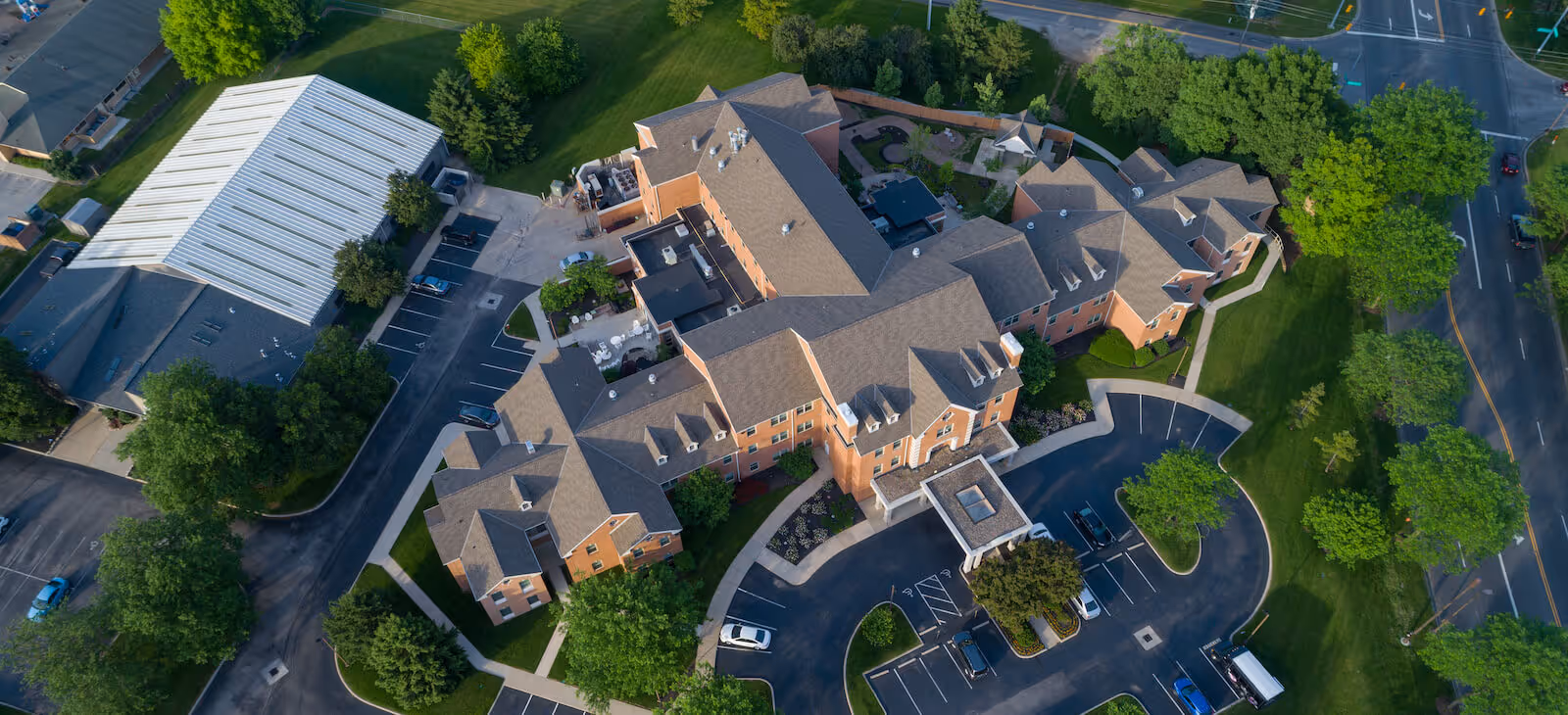 Aerial view of a brick senior living facility with connected buildings, circular driveway, parking lots, and surrounding trees and lawns.