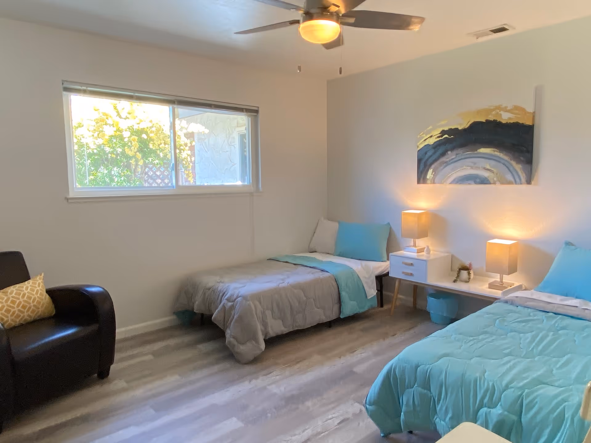 A bright bedroom with two single beds, each with light blue bedding and pillows. Between the beds is a white nightstand with two lit lamps and a small decorative item. A modern abstract painting with gold and blue tones hangs above the nightstand. There is a black armchair with a yellow patterned pillow near a window that lets in natural light. The room has light-colored walls and wood flooring, with a ceiling fan overhead.