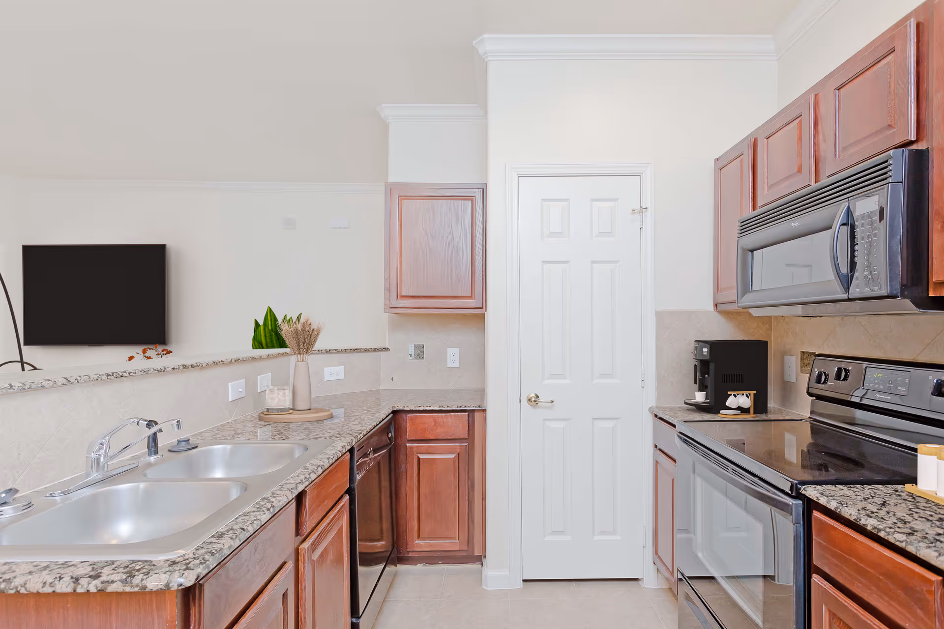 Modern kitchen with granite countertops, wooden cabinets, double sink, stainless steel stove, microwave, dishwasher, and a coffee maker. A white door is visible in the center background.