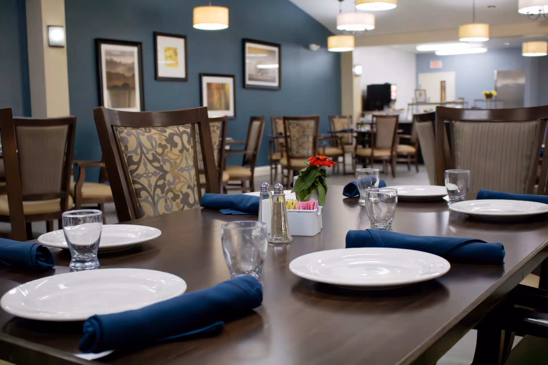 A dining room in an assisted living facility with a long wooden table set with white plates, clear drinking glasses, and blue cloth napkins. The table has a small centerpiece with a red flower and salt and pepper shakers. The room has multiple chairs with patterned upholstery and framed artwork on the blue walls. Ceiling lights illuminate the space.