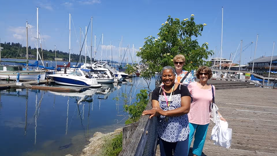 Three women standing on a wooden dock near a marina with several sailboats and motorboats docked in the water. The sky is clear and blue, and there is greenery behind the women.