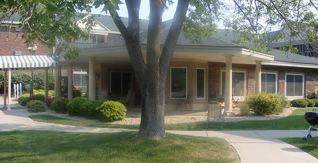 Exterior view of a single-story brick building with a covered entrance, surrounded by green grass, bushes, and trees on a sunny day.
