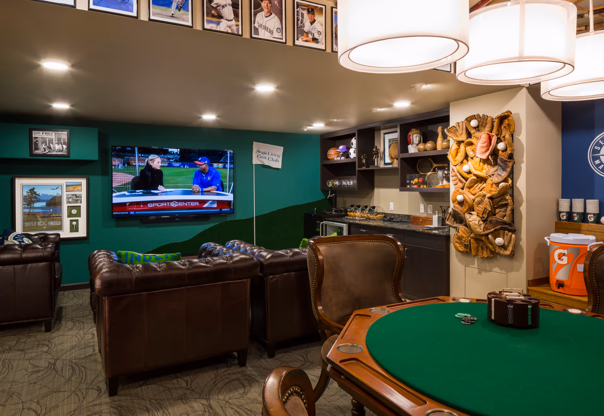A cozy lounge area with brown leather sofas facing a wall-mounted TV showing a sports program. The room has a green accent wall decorated with framed pictures and a small flag labeled 'Aegis Living Golf Club'. There is a poker table with chairs in the foreground, and a wall display featuring numerous baseball gloves and balls. The room also includes a kitchenette area with shelves holding sports memorabilia and a Gatorade cooler with cups stacked nearby. Large hanging lights illuminate the space.