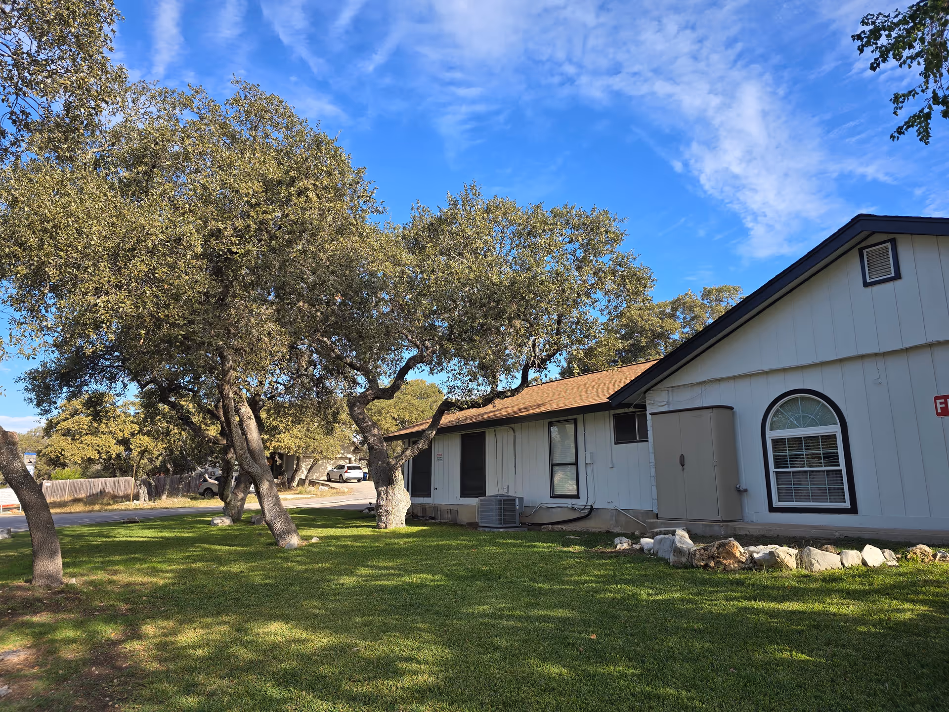 Exterior view of a single-story building with white siding and a brown roof, surrounded by green grass and several large trees under a blue sky with some clouds.