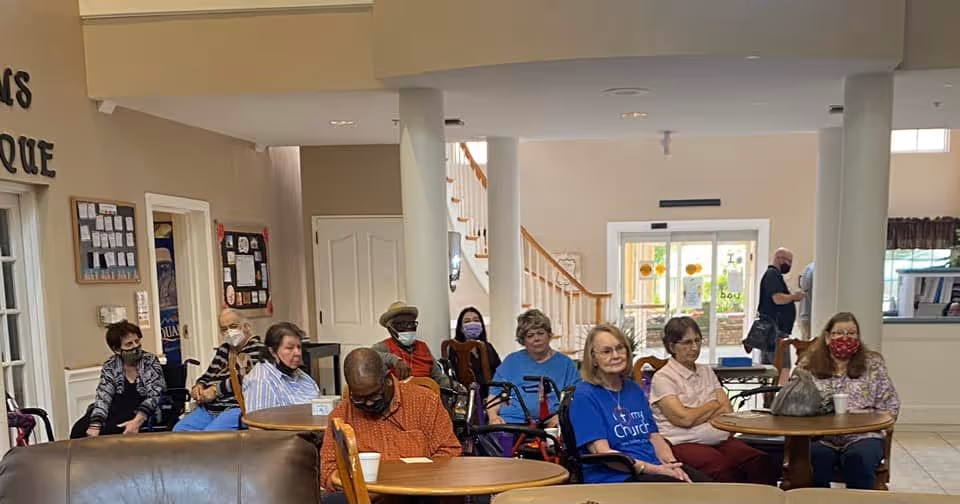 A group of elderly people sitting at round tables in a common area of an assisted living facility. Some individuals are wearing face masks. The room has beige walls, white pillars, and a staircase in the background. There are doors and windows letting in natural light.