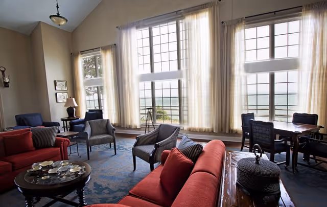 A spacious living room with large windows covered by sheer curtains, offering a view of the ocean outside. The room features red and striped armchairs, a round coffee table with decorative items, a wooden side table with a lamp, and a dining table with chairs near the windows. The room has high ceilings and a blue patterned carpet.
