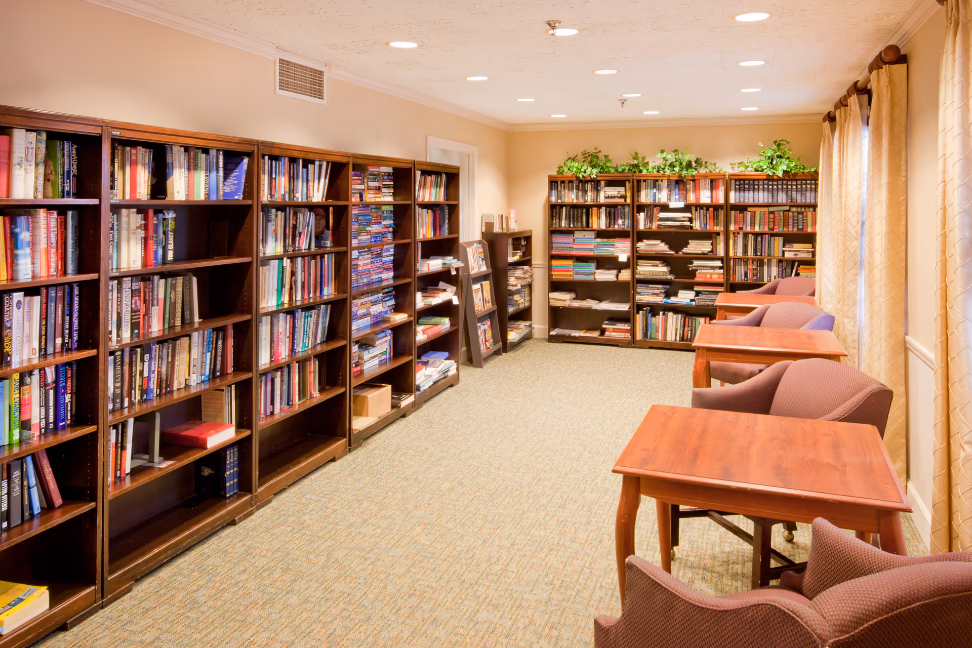 A well-lit library room with multiple wooden bookshelves filled with books along the left and back walls. There are several wooden tables with comfortable upholstered chairs arranged near large windows with beige curtains on the right side. The room has a carpeted floor and recessed ceiling lights, with some green plants placed on top of the bookshelves.