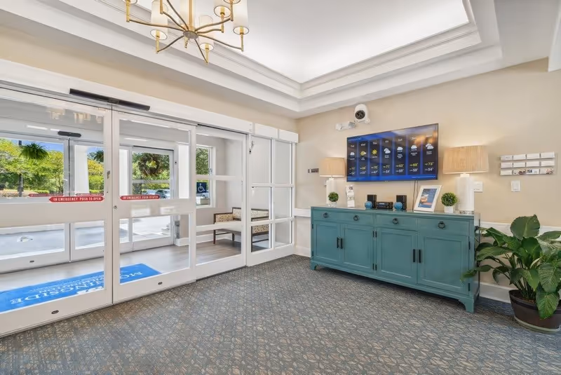 Entrance area of Morningside of Wilmington facility with automatic sliding glass doors, a blue cabinet with decorative items and two lamps, a wall-mounted TV displaying a weather forecast, and a potted plant on the right side.