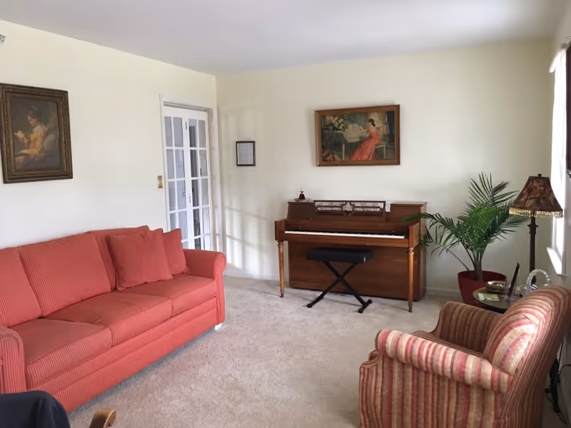 A cozy living room with a red sofa on the left and a striped armchair on the right. In the center against the wall is an upright piano with a black bench. There are two framed paintings on the walls, one above the piano and one above the sofa. A potted plant and a floor lamp with a decorative lampshade are near the window on the right side. The room has light-colored walls and carpeted flooring.