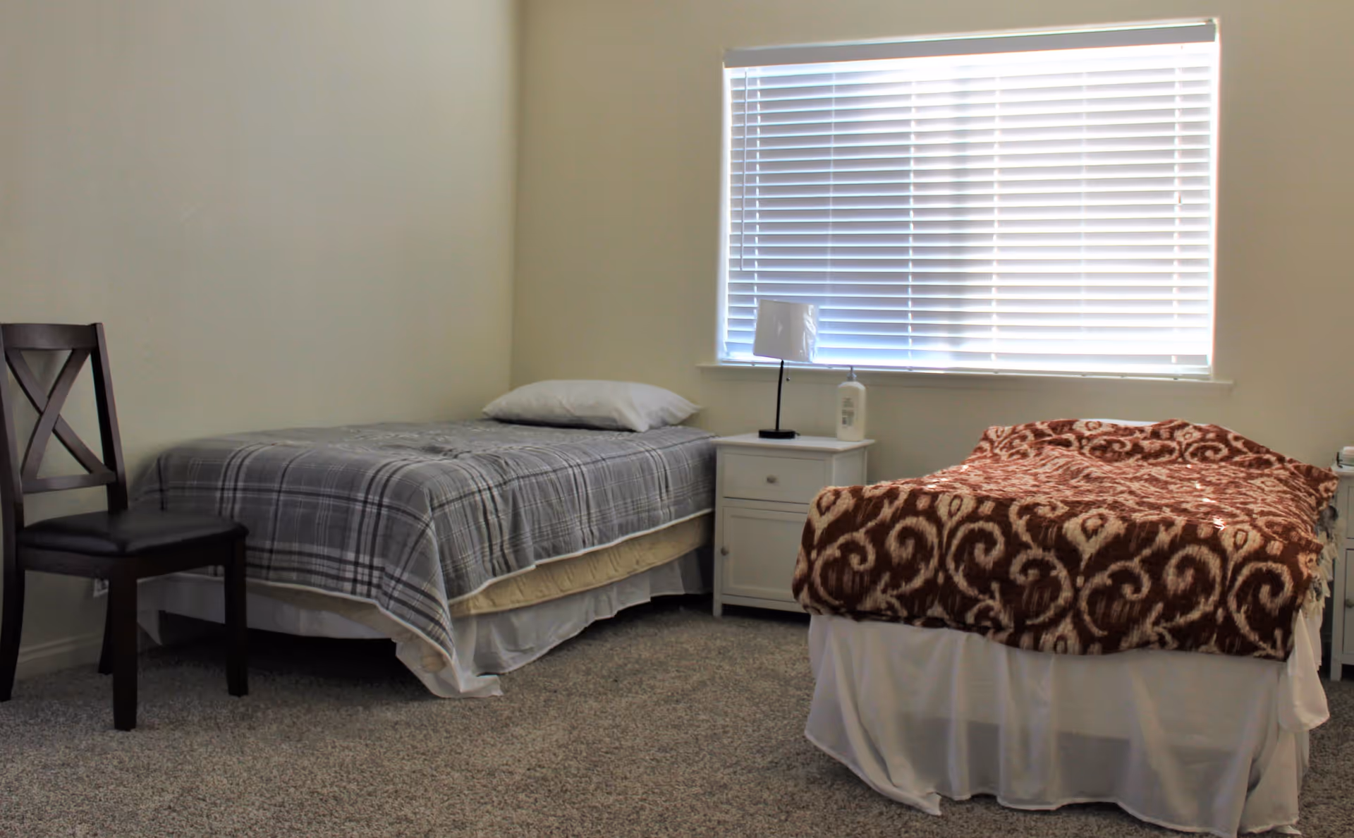 A simple bedroom with two single beds, one with a gray plaid blanket and a white pillow, the other with a brown patterned blanket. Between the beds is a white nightstand with a small lamp and a bottle of lotion. A wooden chair is placed next to the bed on the left. A window with closed blinds is on the wall behind the beds.