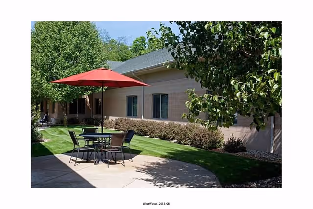 Outdoor patio area at West Woods of Niles featuring a round table with four chairs and a red umbrella, surrounded by green grass, trees, and bushes next to a beige building under a clear blue sky.