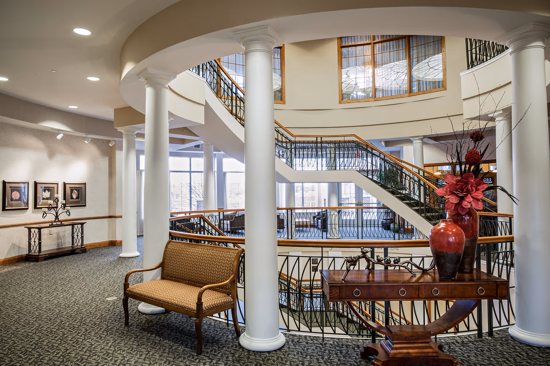 Interior view of a senior living facility with a curved staircase, white columns, and large windows allowing natural light. There is a wooden bench with patterned upholstery and a wooden table with decorative red vases and flowers. The walls have framed artwork and the carpet has a dark patterned design.
