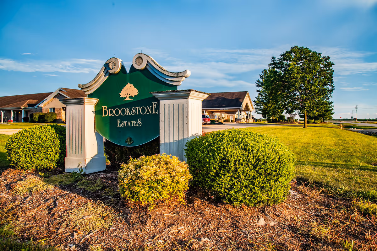 Outdoor view of the entrance sign for Brookstone Estates, surrounded by green bushes and landscaping with a building and trees in the background under a blue sky.