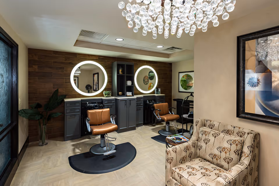 Interior view of a stylish salon area with two brown salon chairs in front of illuminated round mirrors mounted on a wooden accent wall. There is a patterned armchair and a small table with magazines in the foreground, a large decorative chandelier hanging from the ceiling, and framed artwork on the wall.