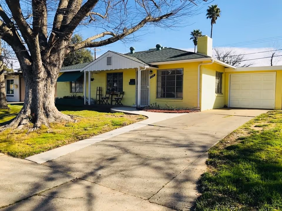 Single-story yellow house with a front porch, attached garage, driveway, and a large tree in the front yard.