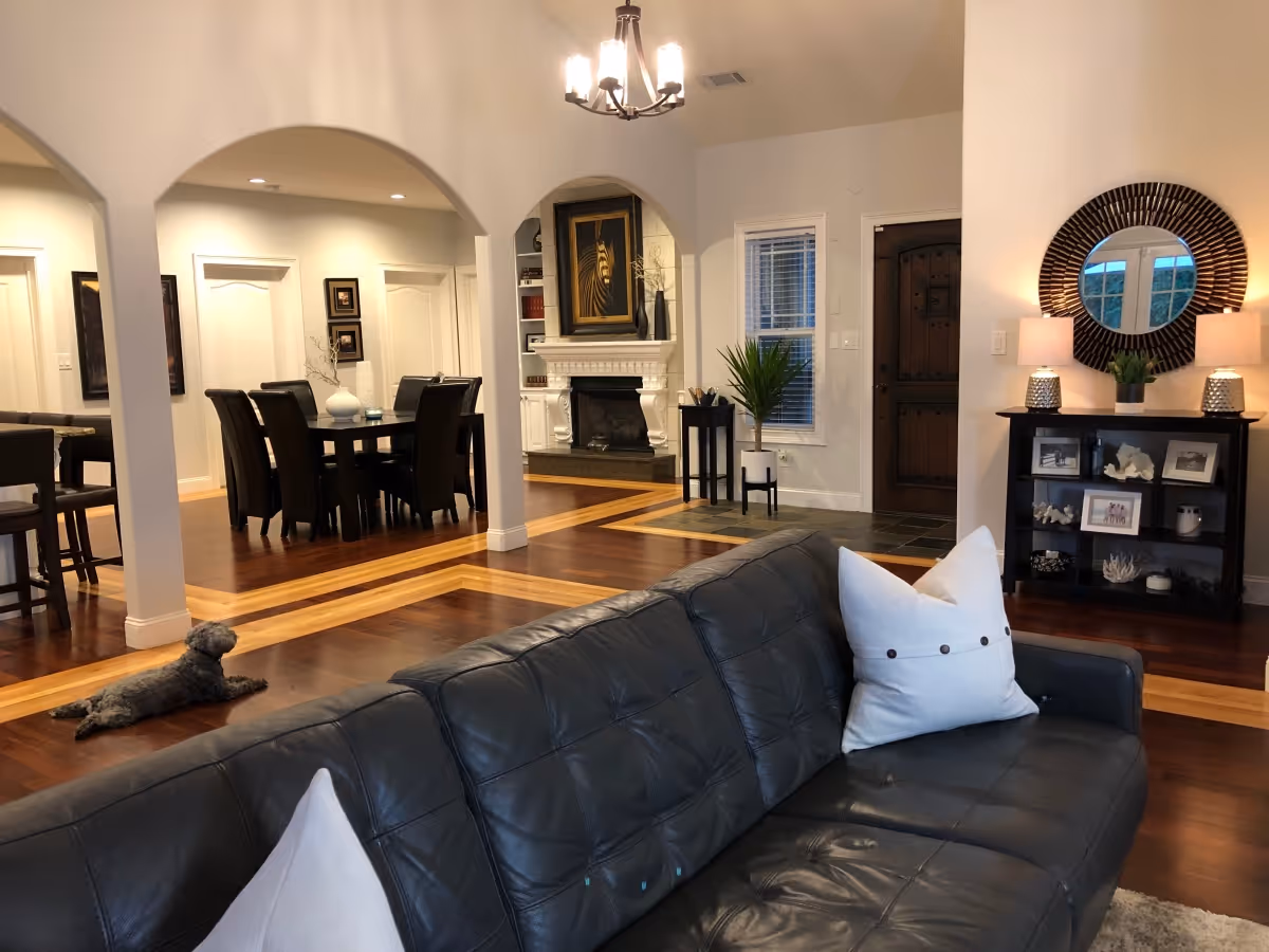 Interior view of a senior living facility showing a spacious living room with a black leather couch adorned with white pillows. In the background, there is a dining area with a dark wood table and chairs, a fireplace with artwork above it, and a wooden front door. The room features hardwood floors with a lighter wood inlay, arched doorways, and decorative elements including a round mirror, lamps, plants, and framed pictures. A small dog is lying on the floor near the dining area.