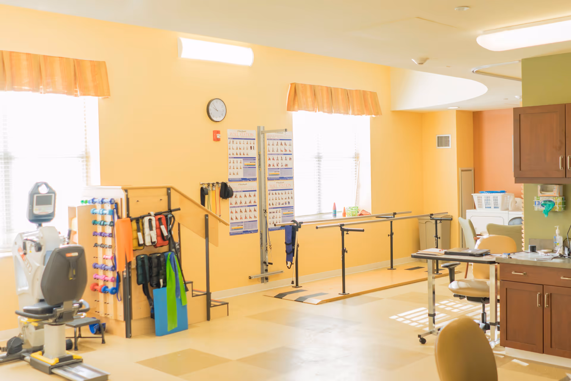 A bright physical therapy room with yellow walls and large windows covered with orange valances. The room contains exercise equipment including a stationary bike, hand weights, resistance bands, parallel bars for walking practice, and a small set of stairs with a handrail. There are instructional posters on the wall and a clock above them. A countertop with cabinets and chairs is visible on the right side.