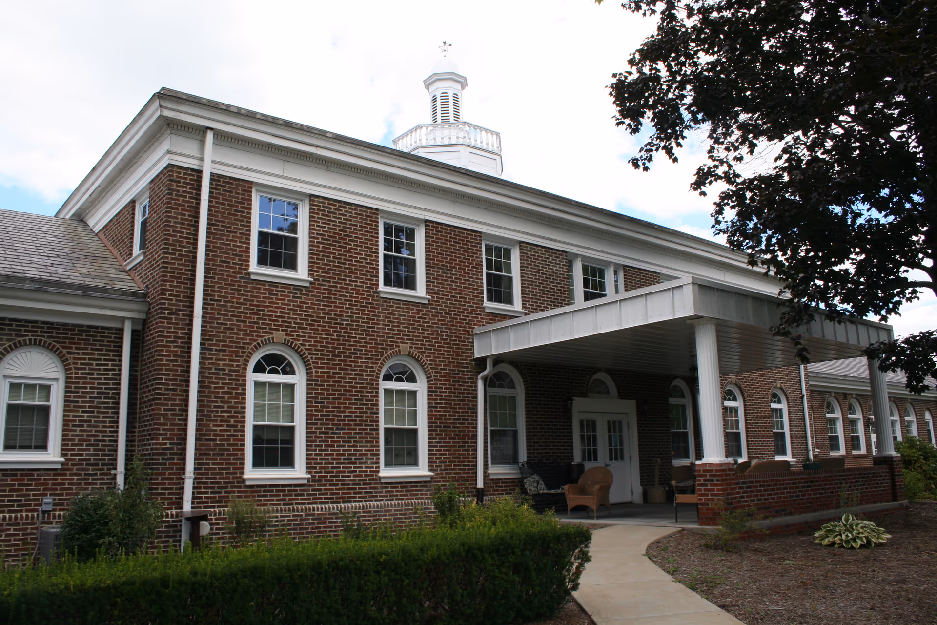 Exterior view of a two-story brick building with white trim and multiple windows. The building has a covered entrance with white columns and outdoor seating. There is a small garden with bushes and a tree nearby.