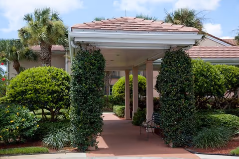 Covered walkway entrance with a tiled roof supported by columns wrapped in greenery, surrounded by well-maintained bushes and palm trees under a partly cloudy sky.
